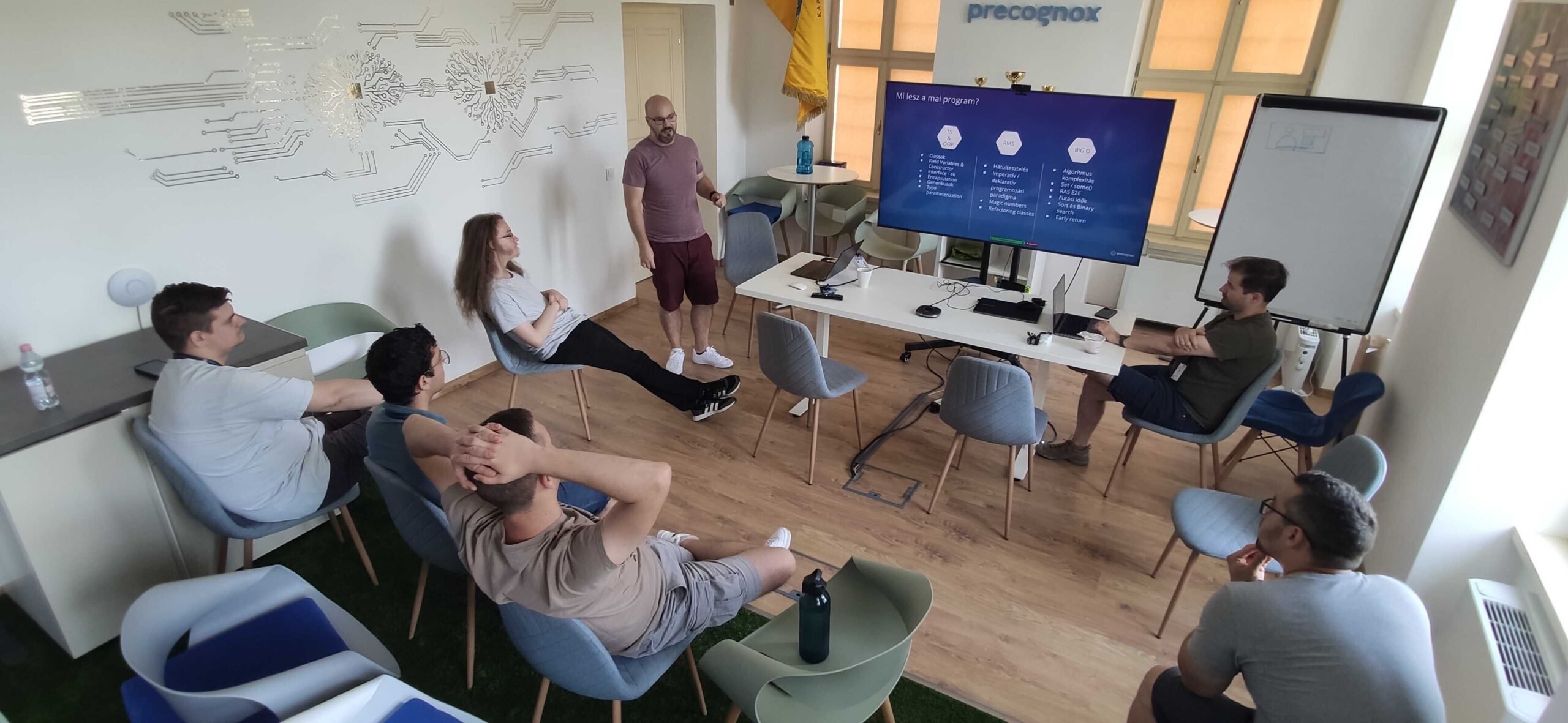 A man is presenting on a big screen for 8 people sitting on chairs.