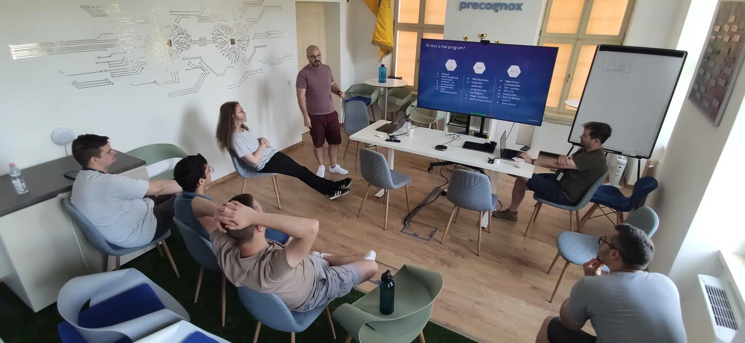 A man is presenting on a big screen for 8 people sitting on chairs.