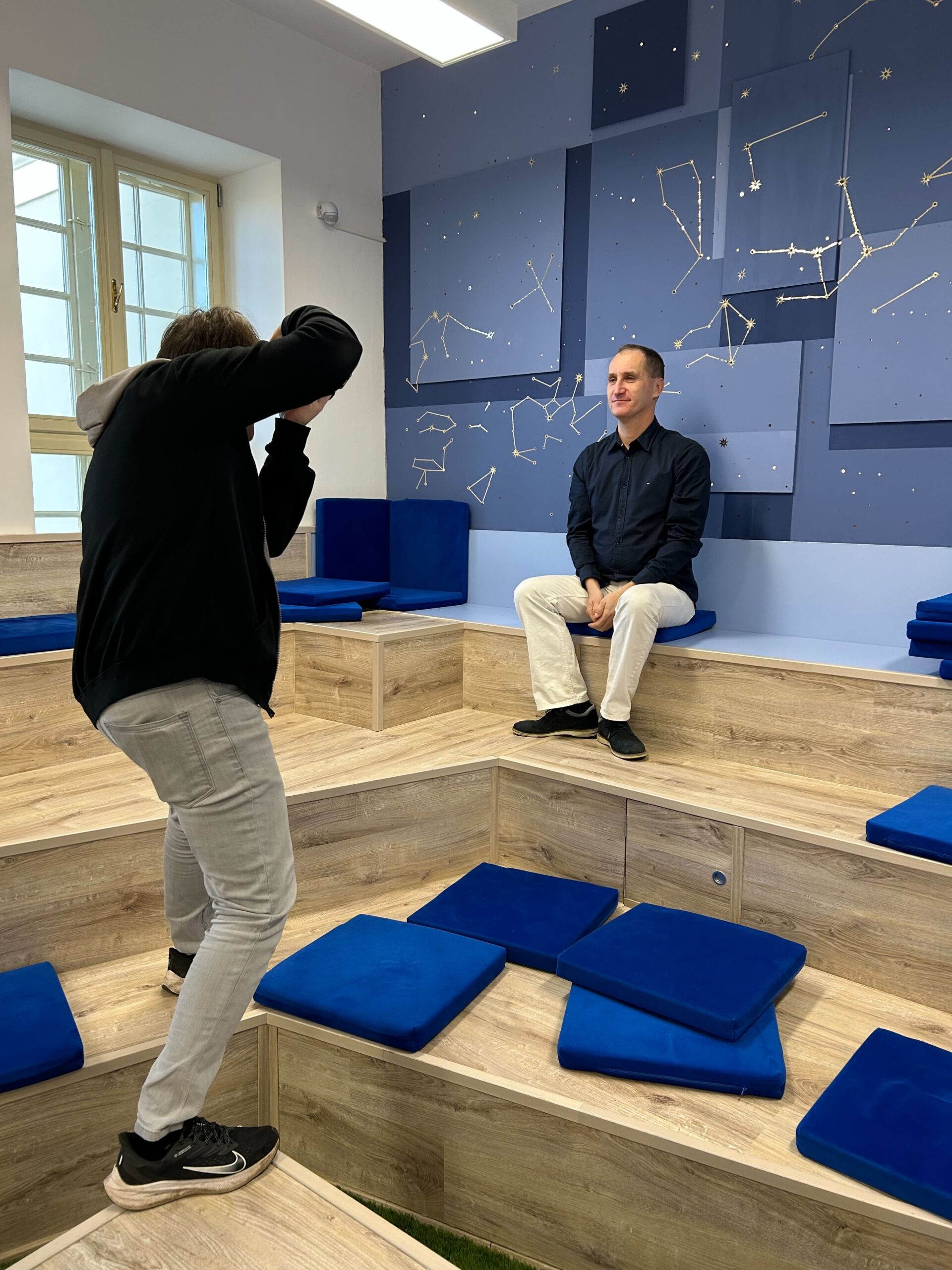 A man sitting in front of a wall decorated with stars. Another man is making a photo of him.